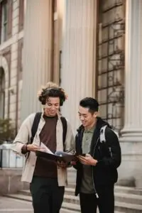 Smiling multiracial classmates in casual wear discussing report together standing near historic university building